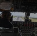 The image shows the interior of an aircraft cockpit from behind, with a pilot seated at the controls during what appears to be a takeoff or landing roll on a wet runway. The cockpit features an older-generation analog instrument panel with numerous gauges, switches, and red-guarded controls. Mountains and green terrain are visible in the background through the windscreen.