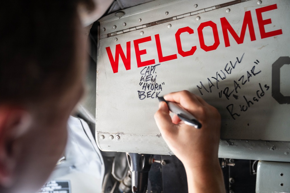A person is writing names on a metal panel of what appears to be a military aircraft. The panel displays a large red 'WELCOME' text along with handwritten names including 'CAPT. KEN "HYDRA" BECK' and 'MAXWELL "RAZAR" RICHARDS'. This appears to be a traditional military welcome or naming ceremony.