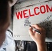 A person is writing names on a metal panel of what appears to be a military aircraft. The panel displays a large red 'WELCOME' text along with handwritten names including 'CAPT. KEN "HYDRA" BECK' and 'MAXWELL "RAZAR" RICHARDS'. This appears to be a traditional military welcome or naming ceremony.