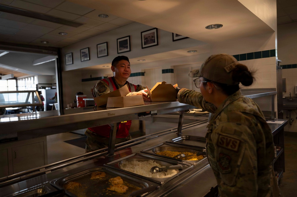 Two military personnel are interacting at a dining facility (DFAC) serving line. One service member wearing a red vest is handing a to-go food container across the stainless steel serving counter to another service member with 'SVS' patch visible on their uniform. The setting appears to be a military base cafeteria with food trays visible in the serving line.