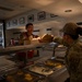 Two military personnel are interacting at a dining facility (DFAC) serving line. One service member wearing a red vest is handing a to-go food container across the stainless steel serving counter to another service member with 'SVS' patch visible on their uniform. The setting appears to be a military base cafeteria with food trays visible in the serving line.
