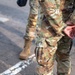 The image shows one or two military personnel wearing OCP (Operational Camouflage Pattern) uniforms and tan combat boots. The foreground figure is holding a rifle (appears to be an M4-style carbine) with an optic and accessory rail attachments at their side, with hands clasped behind their back. The setting appears to be an outdoor paved area, possibly a checkpoint or security post.