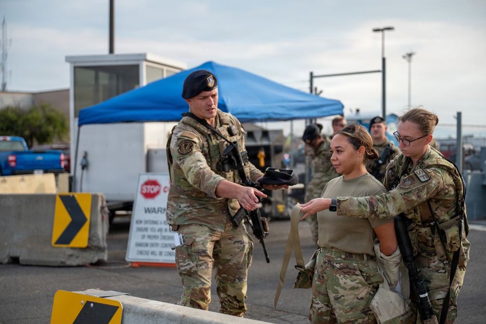 Military personnel in camouflage uniforms are gathered at what appears to be a security checkpoint on a military installation. A male soldier wearing a black beret is interacting with two female soldiers, appearing to conduct training or a briefing near a controlled entry point. A blue canopy tent, concrete barriers, and a STOP sign are visible in the background.