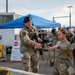 Military personnel in camouflage uniforms are gathered at what appears to be a security checkpoint on a military installation. A male soldier wearing a black beret is interacting with two female soldiers, appearing to conduct training or a briefing near a controlled entry point. A blue canopy tent, concrete barriers, and a STOP sign are visible in the background.