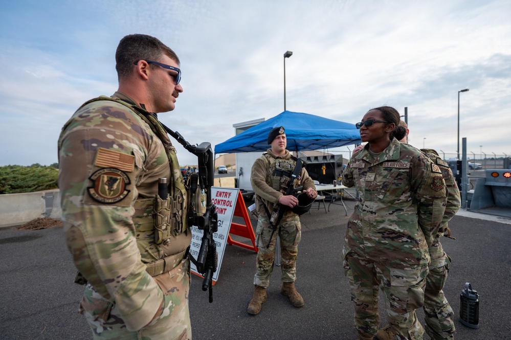 Three U.S. Air Force personnel in OCP (Operational Camouflage Pattern) uniforms are gathered at a military Entry Control Point. Two appear to be armed security forces personnel equipped with tactical gear and rifles, while the third displays 'U.S. AIR FORCE' tape on their uniform. They appear to be engaged in a briefing or conversation near a blue canopy tent.