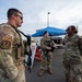 Three U.S. Air Force personnel in OCP (Operational Camouflage Pattern) uniforms are gathered at a military Entry Control Point. Two appear to be armed security forces personnel equipped with tactical gear and rifles, while the third displays 'U.S. AIR FORCE' tape on their uniform. They appear to be engaged in a briefing or conversation near a blue canopy tent.