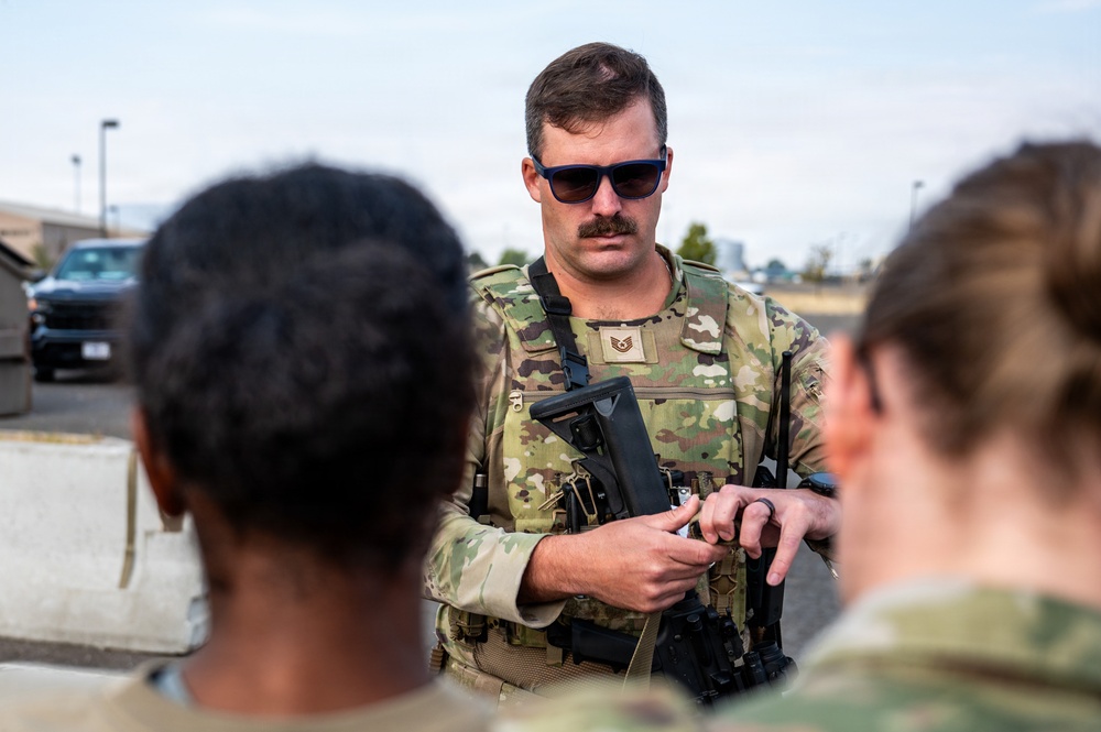 A U.S. Air Force Technical Sergeant in Operational Camouflage Pattern (OCP) uniform is conducting what appears to be a weapons briefing or instruction session outdoors. He is wearing a tactical vest, sunglasses, and holding a firearm while addressing at least two other personnel whose backs are visible in the foreground. The setting appears to be a military installation or base.