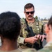 A U.S. Air Force Technical Sergeant in Operational Camouflage Pattern (OCP) uniform is conducting what appears to be a weapons briefing or instruction session outdoors. He is wearing a tactical vest, sunglasses, and holding a firearm while addressing at least two other personnel whose backs are visible in the foreground. The setting appears to be a military installation or base.