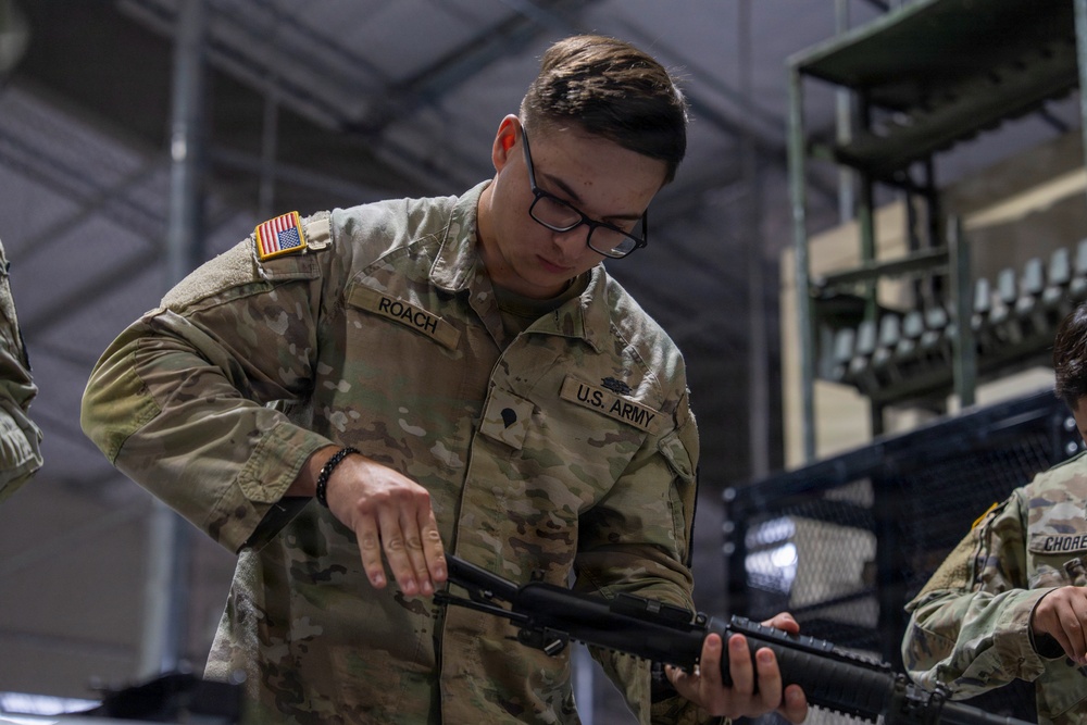 A U.S. Army soldier named Roach is performing maintenance or inspection on a rifle in what appears to be an armory or equipment facility. The soldier is wearing the standard Operational Camouflage Pattern (OCP) uniform with a U.S. flag patch on the shoulder. A second soldier, partially visible on the right with the name tape 'CHORE', is also present in the same environment.