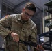 A U.S. Army soldier named Roach is performing maintenance or inspection on a rifle in what appears to be an armory or equipment facility. The soldier is wearing the standard Operational Camouflage Pattern (OCP) uniform with a U.S. flag patch on the shoulder. A second soldier, partially visible on the right with the name tape 'CHORE', is also present in the same environment.