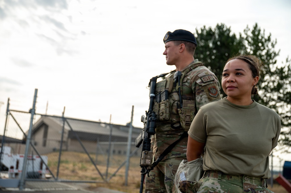 Two U.S. Air Force Security Forces (SF) personnel are standing outdoors at what appears to be a military installation. The male airman in the background is wearing a black beret and carrying a rifle, while the female airman in the foreground is in a standard OCP uniform. Both appear to be on duty, surveying their surroundings near a fenced perimeter.