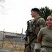 Two U.S. Air Force Security Forces (SF) personnel are standing outdoors at what appears to be a military installation. The male airman in the background is wearing a black beret and carrying a rifle, while the female airman in the foreground is in a standard OCP uniform. Both appear to be on duty, surveying their surroundings near a fenced perimeter.