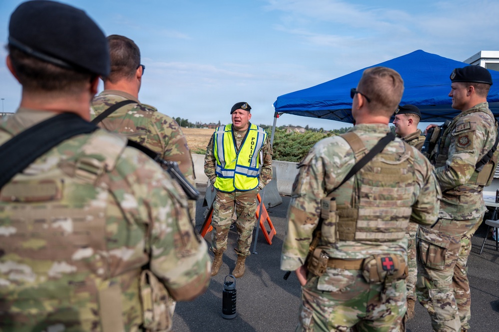 A military police officer wearing a yellow high-visibility vest labeled 'POLICE' is briefing a group of soldiers in OCP (Operational Camouflage Pattern) uniforms outdoors. The personnel appear to be conducting a security checkpoint or access control point operation, with concrete barriers and a blue canopy tent visible in the background. The setting appears to be a military installation or controlled entry point.
