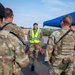 A military police officer wearing a yellow high-visibility vest labeled 'POLICE' is briefing a group of soldiers in OCP (Operational Camouflage Pattern) uniforms outdoors. The personnel appear to be conducting a security checkpoint or access control point operation, with concrete barriers and a blue canopy tent visible in the background. The setting appears to be a military installation or controlled entry point.