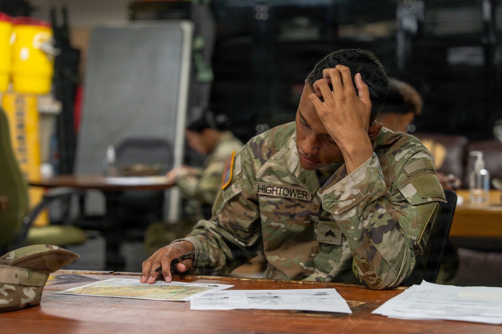 A U.S. Army soldier named Hightower, wearing Operational Camouflage Pattern (OCP) uniform, sits at a table studying documents and maps with one hand pressed against his head in a concentrated or stressed posture. The setting appears to be a military classroom or operations room with other soldiers visible in the background. Documents, printed materials, and what appears to be a military map are spread on the table in front of him.