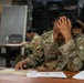 A U.S. Army soldier named Hightower, wearing Operational Camouflage Pattern (OCP) uniform, sits at a table studying documents and maps with one hand pressed against his head in a concentrated or stressed posture. The setting appears to be a military classroom or operations room with other soldiers visible in the background. Documents, printed materials, and what appears to be a military map are spread on the table in front of him.