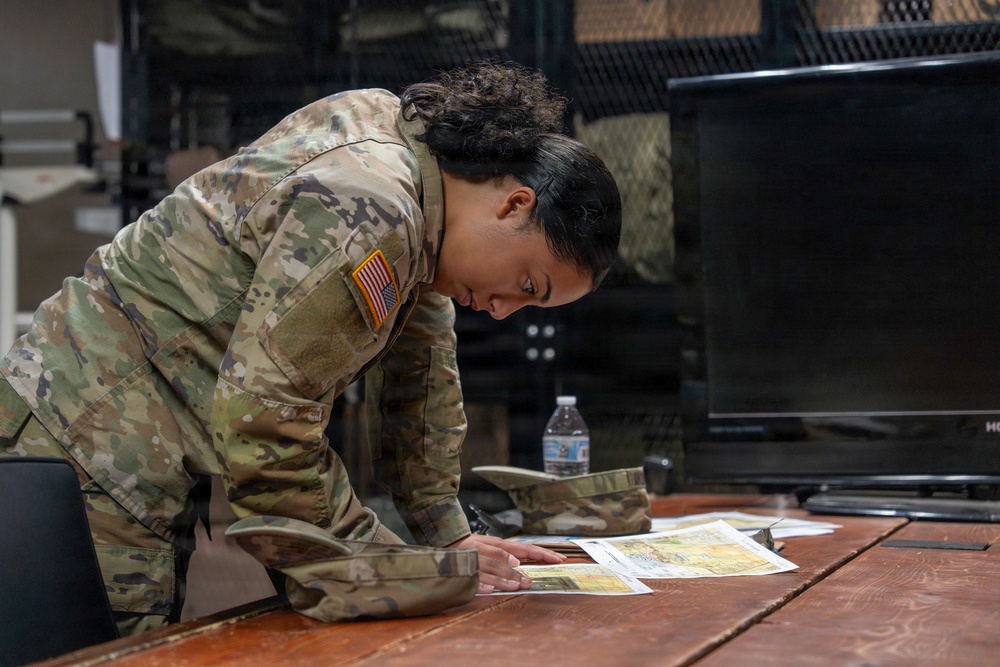 A U.S. Army soldier in Operational Camouflage Pattern (OCP) uniform is leaning over a wooden table, examining what appear to be maps or printed documents. The soldier wears a U.S. flag patch on the right sleeve, indicating active U.S. military affiliation. The setting appears to be an indoor military operations or briefing room.