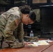 A U.S. Army soldier in Operational Camouflage Pattern (OCP) uniform is leaning over a wooden table, examining what appear to be maps or printed documents. The soldier wears a U.S. flag patch on the right sleeve, indicating active U.S. military affiliation. The setting appears to be an indoor military operations or briefing room.