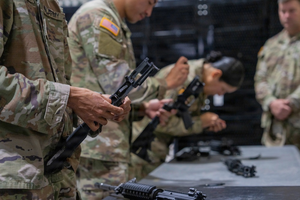 U.S. Army soldiers in MultiCam/OCP camouflage uniforms are conducting a weapons familiarization or maintenance training session indoors. Multiple personnel are handling disassembled firearms at a table, suggesting a field stripping or weapons inspection exercise. The setting appears to be an indoor training facility or armory.