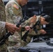 U.S. Army soldiers in MultiCam/OCP camouflage uniforms are conducting a weapons familiarization or maintenance training session indoors. Multiple personnel are handling disassembled firearms at a table, suggesting a field stripping or weapons inspection exercise. The setting appears to be an indoor training facility or armory.