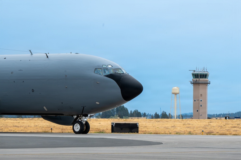 A large military aircraft, painted in dark gray/charcoal livery, is taxiing on a flight line at a military airbase. The nose and forward fuselage section dominate the foreground, with the nose gear visible on the tarmac. In the background, an air traffic control tower and a water tower are visible alongside a treeline under an overcast sky.