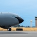 A large military aircraft, painted in dark gray/charcoal livery, is taxiing on a flight line at a military airbase. The nose and forward fuselage section dominate the foreground, with the nose gear visible on the tarmac. In the background, an air traffic control tower and a water tower are visible alongside a treeline under an overcast sky.