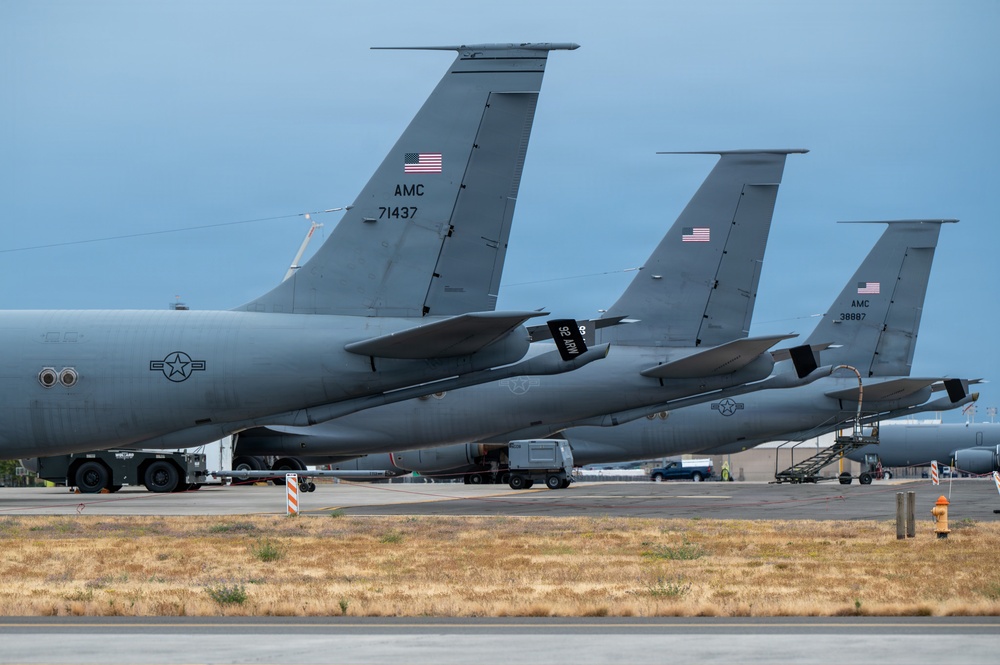 The image shows multiple U.S. Air Force AMC (Air Mobility Command) aircraft parked on a flight line, viewed from the rear showing their tail sections. At least two tail numbers are visible: 71437 and 38887, consistent with KC-135 Stratotanker aircraft. The aircraft are painted in standard gray military livery and display American flags on their vertical stabilizers.
