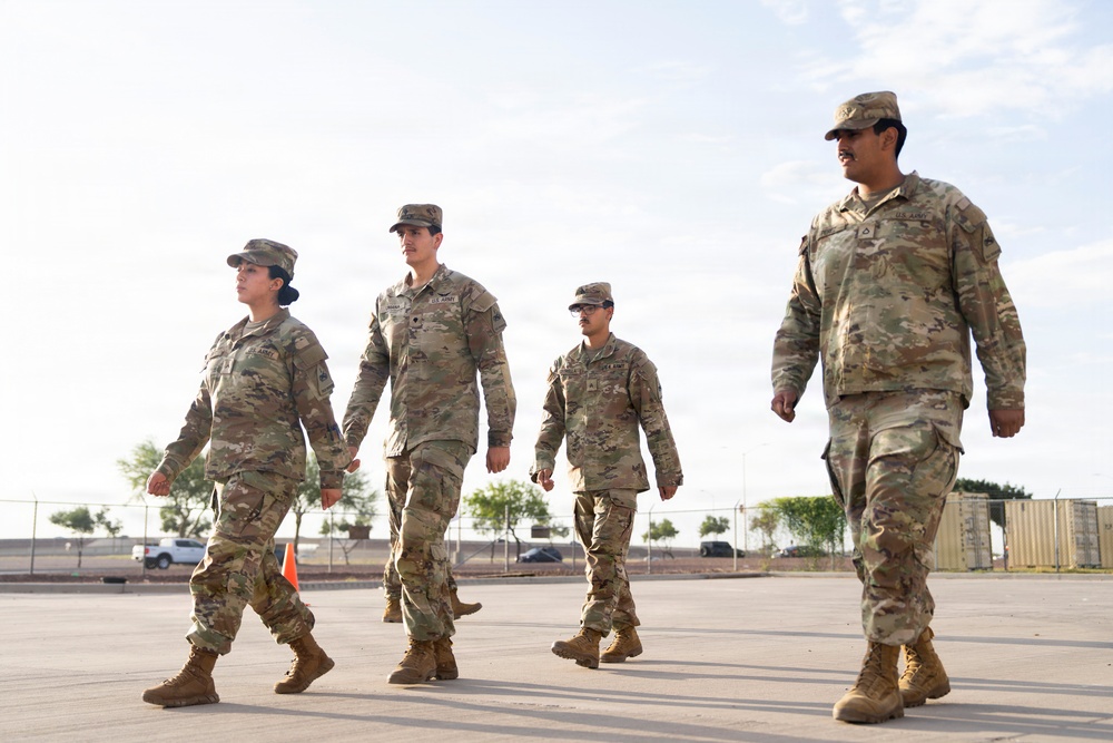 Four U.S. Army soldiers in Operational Camouflage Pattern (OCP) uniforms are walking across a paved surface at what appears to be a military installation or base. The group consists of three soldiers walking together on the left and one soldier slightly ahead on the right. The setting appears to be an outdoor military compound with fencing, vehicles, and storage containers visible in the background.
