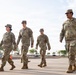 Four U.S. Army soldiers in Operational Camouflage Pattern (OCP) uniforms are walking across a paved surface at what appears to be a military installation or base. The group consists of three soldiers walking together on the left and one soldier slightly ahead on the right. The setting appears to be an outdoor military compound with fencing, vehicles, and storage containers visible in the background.