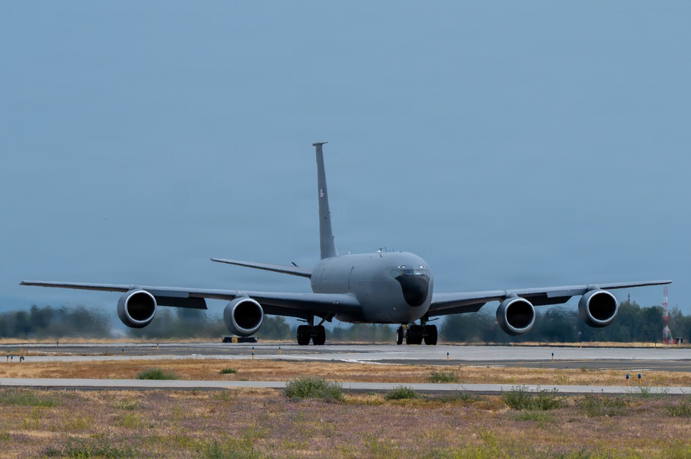 A large four-engine military jet aircraft is taxiing on a runway, viewed from a direct front/nose-on perspective. The aircraft is painted in an overall gray scheme consistent with U.S. Air Force markings, with swept wings and four podded engines. The setting appears to be a military airfield with dry grass and trees visible in the background.