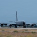 A large four-engine military jet aircraft is taxiing on a runway, viewed from a direct front/nose-on perspective. The aircraft is painted in an overall gray scheme consistent with U.S. Air Force markings, with swept wings and four podded engines. The setting appears to be a military airfield with dry grass and trees visible in the background.