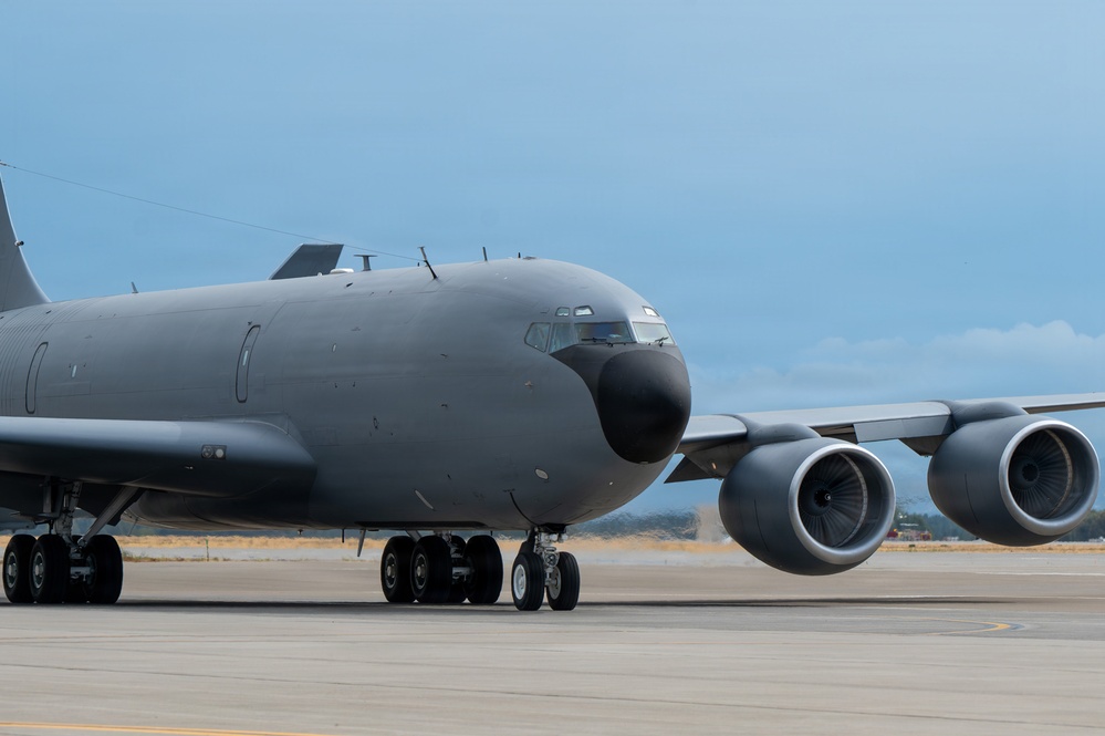A large military aircraft, consistent with a KC-135 Stratotanker or similar tanker/transport variant, is photographed on a tarmac in a low-angle, close-up perspective. The aircraft is painted in an overall dark gray military finish with no visible markings or insignia. Two turbofan engines are prominently visible under the swept wing on the right side of the frame.