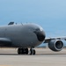 A large military aircraft, consistent with a KC-135 Stratotanker or similar tanker/transport variant, is photographed on a tarmac in a low-angle, close-up perspective. The aircraft is painted in an overall dark gray military finish with no visible markings or insignia. Two turbofan engines are prominently visible under the swept wing on the right side of the frame.
