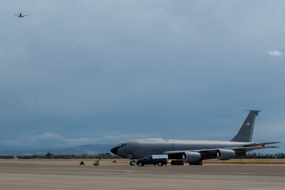 A large military tanker aircraft, consistent with a KC-135 Stratotanker, is parked on a flight line ramp with ground support vehicles and personnel nearby. A second aircraft is visible in the background flying at altitude under overcast skies. Mountains or hills are faintly visible on the horizon.