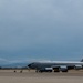 A large military tanker aircraft, consistent with a KC-135 Stratotanker, is parked on a flight line ramp with ground support vehicles and personnel nearby. A second aircraft is visible in the background flying at altitude under overcast skies. Mountains or hills are faintly visible on the horizon.