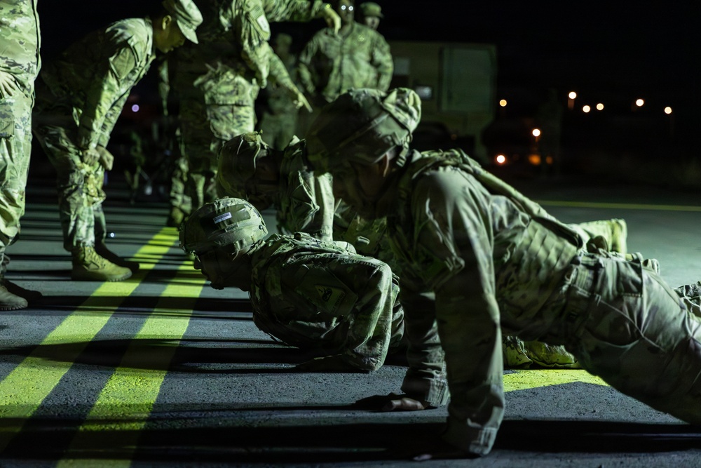 Military personnel in camouflage uniforms are performing push-ups on a tarmac or paved surface at night. The scene appears to be a physical training or disciplinary exercise, with some soldiers standing and observing while others are in the push-up position. The setting is illuminated by artificial lighting against a dark nighttime background.
