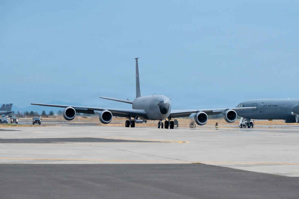 The image shows U.S. Air Force military aircraft taxiing on a flight line at what appears to be a military airbase. The primary aircraft in the foreground is a large gray jet, consistent with a KC-135 Stratotanker aerial refueling aircraft, viewed from the front. A second aircraft marked 'U.S. AIR FORCE' is visible to the right, along with ground support vehicles in the background.