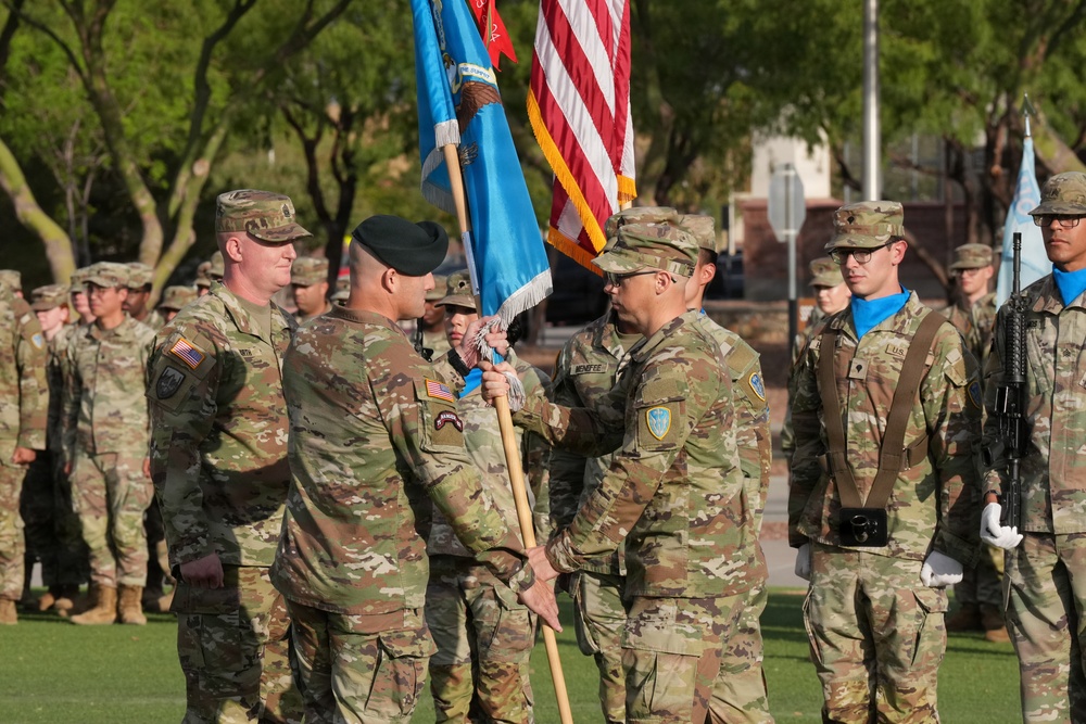 A formal U.S. Army change of command ceremony is taking place outdoors on a parade field. Two senior officers are exchanging a unit guidon (flag), which is the central symbolic act of a change of command. Soldiers in formation stand at attention in the background, with an American flag and unit colors also visible.