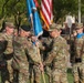 A formal U.S. Army change of command ceremony is taking place outdoors on a parade field. Two senior officers are exchanging a unit guidon (flag), which is the central symbolic act of a change of command. Soldiers in formation stand at attention in the background, with an American flag and unit colors also visible.