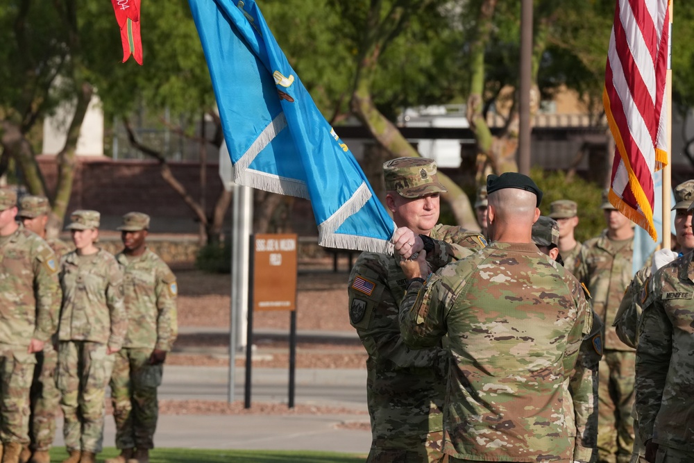A U.S. Army military ceremony is taking place outdoors, likely a change of command or activation ceremony. Two soldiers in the foreground are engaged in a flag or guidon transfer, with one soldier holding a light blue unit flag with silver fringe. A formation of soldiers in OCP (Operational Camouflage Pattern) uniforms stands at attention in the background.