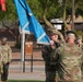 A U.S. Army military ceremony is taking place outdoors, likely a change of command or activation ceremony. Two soldiers in the foreground are engaged in a flag or guidon transfer, with one soldier holding a light blue unit flag with silver fringe. A formation of soldiers in OCP (Operational Camouflage Pattern) uniforms stands at attention in the background.