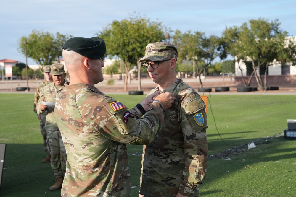 A military ceremony is taking place outdoors on a grass field, where one soldier wearing a green beret is pinning a medal or insignia onto another soldier's uniform. The recipient soldier wears glasses and displays a yellow/blue shoulder patch, while the presenting soldier has a Ranger tab and U.S. flag patch visible on his sleeve. Additional soldiers are visible in the background observing the ceremony.