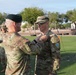 A military ceremony is taking place outdoors on a grass field, where one soldier wearing a green beret is pinning a medal or insignia onto another soldier's uniform. The recipient soldier wears glasses and displays a yellow/blue shoulder patch, while the presenting soldier has a Ranger tab and U.S. flag patch visible on his sleeve. Additional soldiers are visible in the background observing the ceremony.
