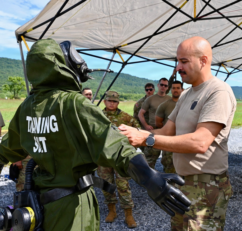 Military personnel are conducting a CBRN (Chemical, Biological, Radiological, Nuclear) training exercise outdoors. A soldier in a green protective suit labeled 'TRAINING SUIT' with a full-face respirator mask is being inspected or assisted by a bald soldier in a tan t-shirt and multicam pants. A group of uniformed soldiers observes the exercise under a canopy tent in a field setting with green hills in the background.