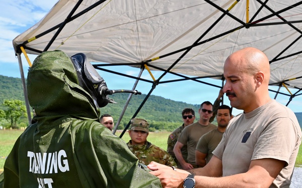 Military personnel are conducting a CBRN (Chemical, Biological, Radiological, Nuclear) training exercise outdoors. A soldier in a green protective suit labeled 'TRAINING SUIT' with a full-face respirator mask is being inspected or assisted by a bald soldier in a tan t-shirt and multicam pants. A group of uniformed soldiers observes the exercise under a canopy tent in a field setting with green hills in the background.