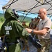 Military personnel are conducting a CBRN (Chemical, Biological, Radiological, Nuclear) training exercise outdoors. A soldier in a green protective suit labeled 'TRAINING SUIT' with a full-face respirator mask is being inspected or assisted by a bald soldier in a tan t-shirt and multicam pants. A group of uniformed soldiers observes the exercise under a canopy tent in a field setting with green hills in the background.