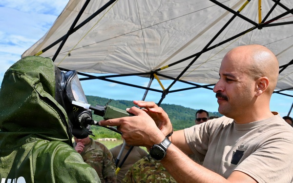 A military training exercise is taking place outdoors under a white canopy tent. A soldier in a tan t-shirt and military pants is adjusting or inspecting the gas mask/face shield of another person wearing a green CBRN (Chemical, Biological, Radiological, Nuclear) protective suit labeled 'TRAINING SUIT'. Additional military personnel in camouflage uniforms are visible in the background.