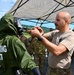 A military training exercise is taking place outdoors under a white canopy tent. A soldier in a tan t-shirt and military pants is adjusting or inspecting the gas mask/face shield of another person wearing a green CBRN (Chemical, Biological, Radiological, Nuclear) protective suit labeled 'TRAINING SUIT'. Additional military personnel in camouflage uniforms are visible in the background.