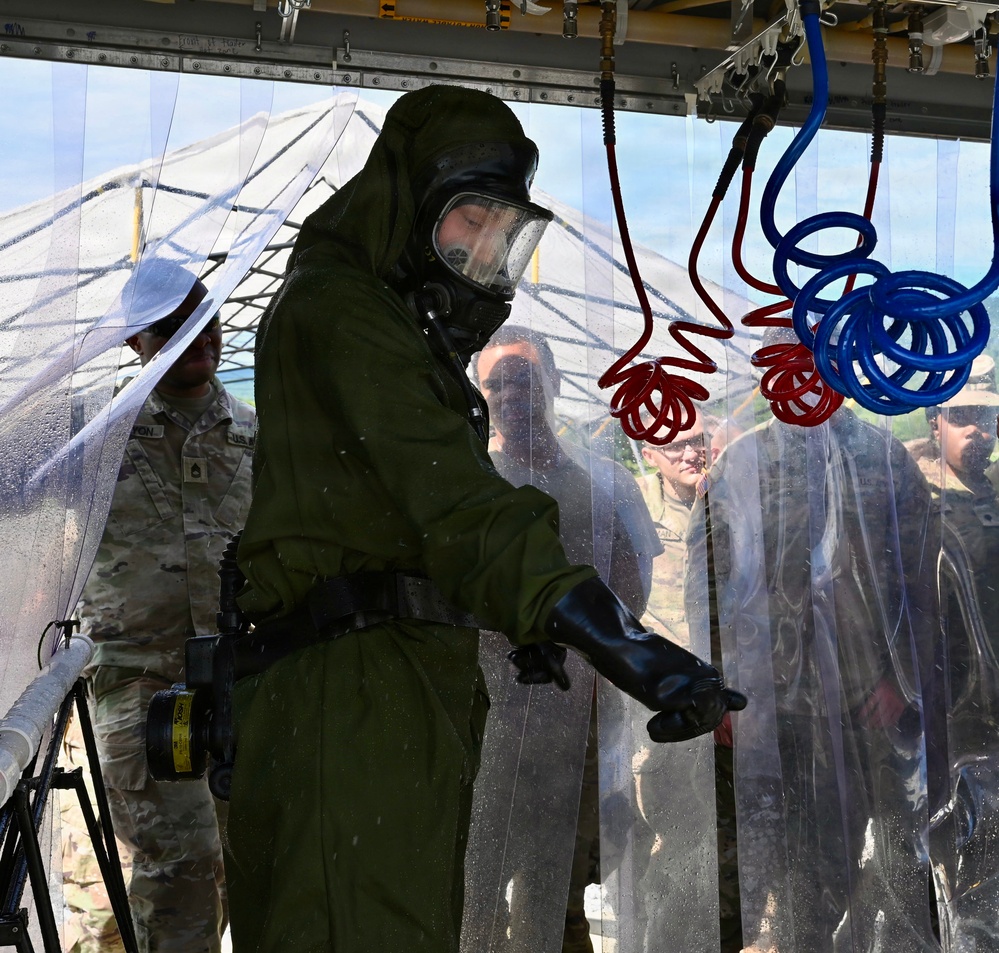 A military personnel wearing full CBRN (Chemical, Biological, Radiological, and Nuclear) protective gear, including a gas mask and olive-green hazmat suit with black rubber gloves, is undergoing a decontamination procedure inside a transparent plastic curtain enclosure. Red and blue coiled hoses are suspended overhead, likely part of a decontamination wash system. Several other military personnel in standard camouflage uniforms observe from outside the enclosure.