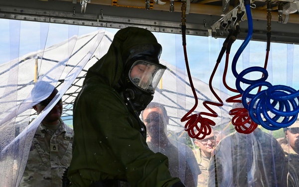 A military personnel wearing full CBRN (Chemical, Biological, Radiological, and Nuclear) protective gear, including a gas mask and olive-green hazmat suit with black rubber gloves, is undergoing a decontamination procedure inside a transparent plastic curtain enclosure. Red and blue coiled hoses are suspended overhead, likely part of a decontamination wash system. Several other military personnel in standard camouflage uniforms observe from outside the enclosure.